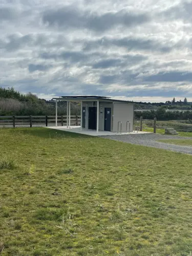 A modern public toilet block sits beside a gravel walkway in an open grassy reserve. The small building has a covered entrance, two doorways, bike stands, and fencing nearby. The surrounding landscape includes wetland vegetation and distant trees beneath a dramatic cloudy sky.
