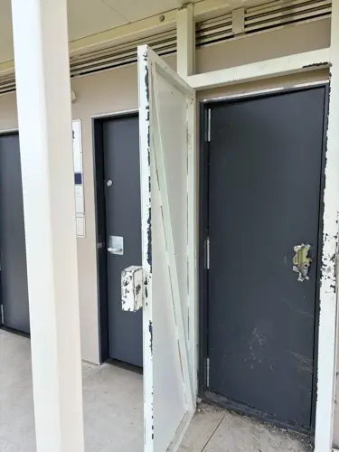 Close-up of a public toilet block showing a damaged entrance gate and a toilet door with the handle removed. The image highlights visible vandalism, including chipped paint, scratches, and damage to fixtures on the exterior of the building.
